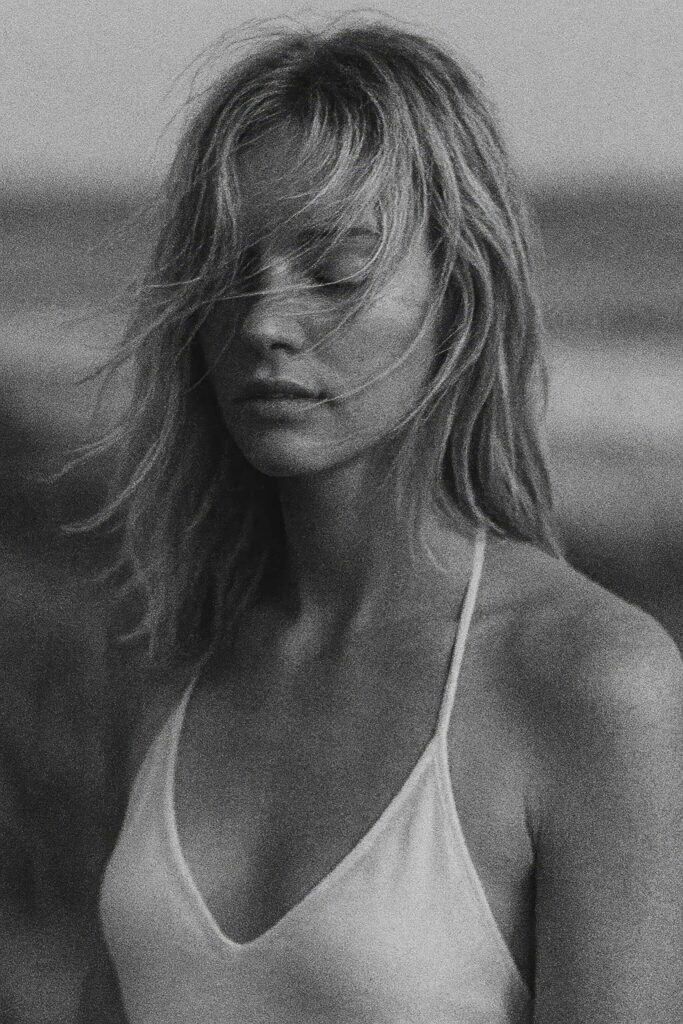 Black and white portrait of a woman with windswept hair, wearing a white camisole top. She stands with her eyes closed, evoking a quiet and emotional moment by the coast. The soft grain and natural light create a moody, cinematic feel. Ideal for editorial, lifestyle, or artistic visual storytelling. Created with the assistance of AI tools.